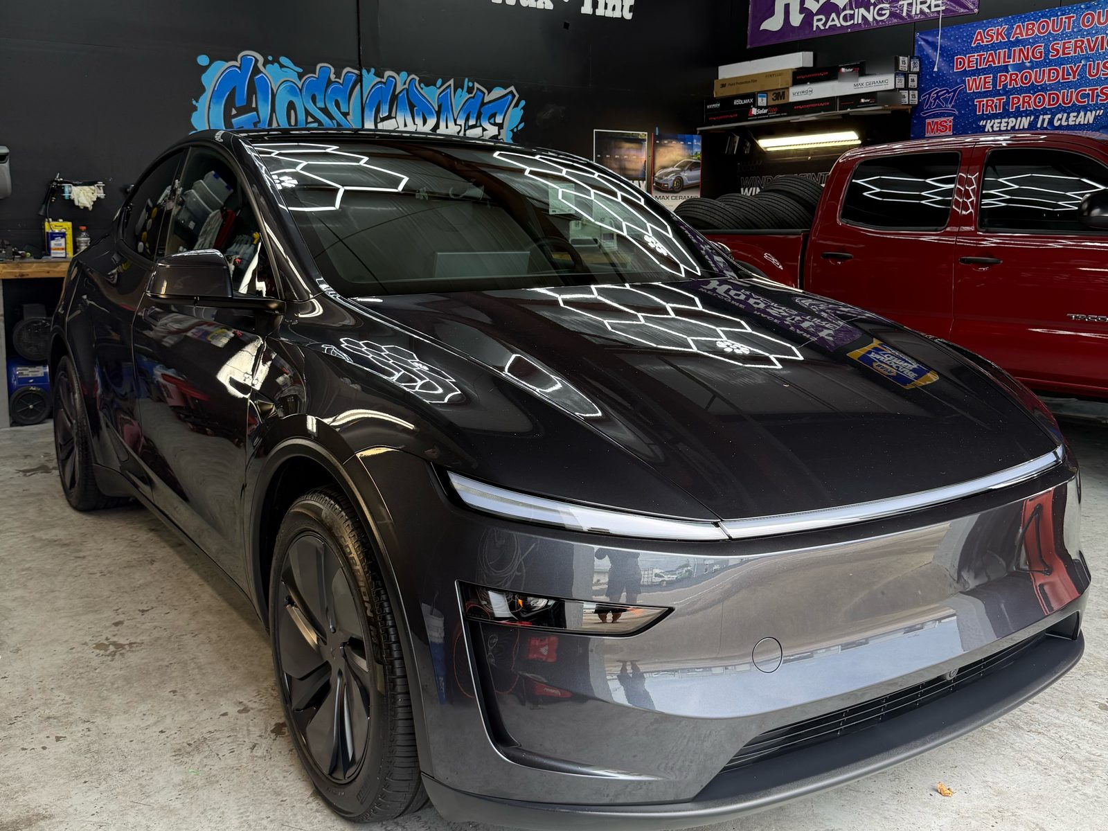 Gray Tesla Model Y front three-quarter view after ceramic coating, parked beside a red truck in our shop